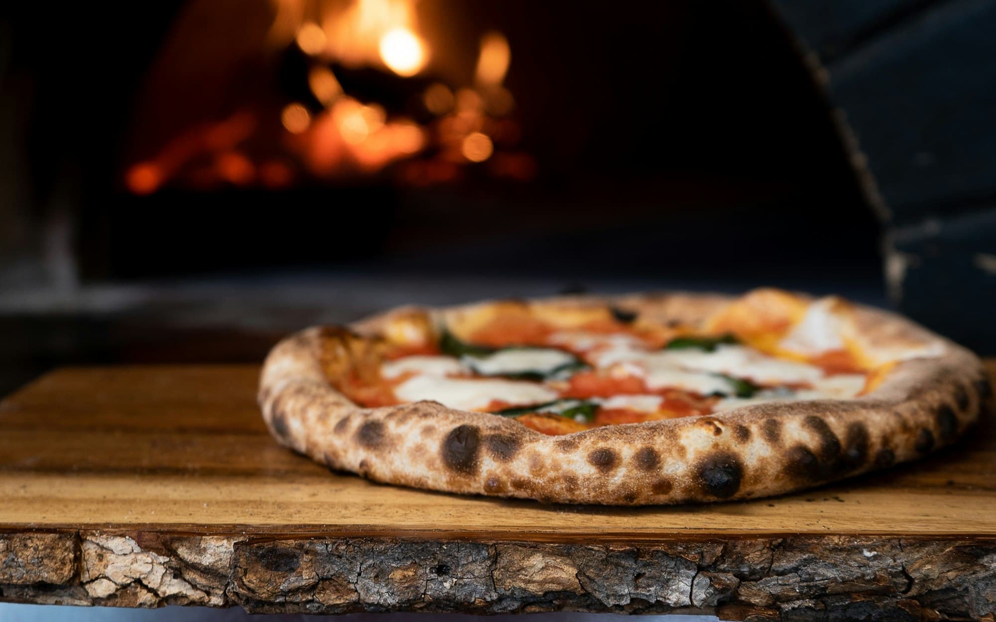 A wood-fired pizza resting on a board in front of a burning oven